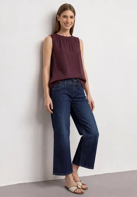 Frau in lila Top, weiten blauen Jeans und Sandalen steht an der Wand und lächelt.