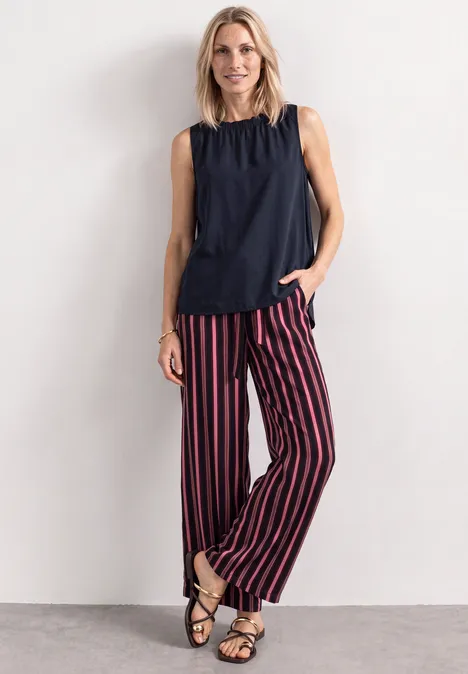 Frau in schwarzem Top, rot-schwarz gestreiften Hosen und Sandalen posiert vor heller Wand.