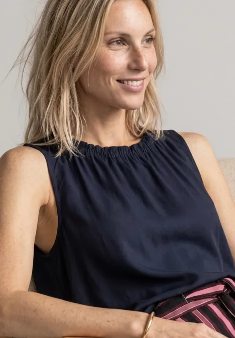 Lächelnde blonde Frau in ärmellosem dunkelblauen Top vor neutralem Hintergrund.