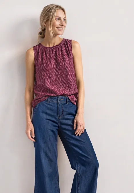 Femme portant un top sans manches rouge et noir et un jean large bleu sur fond blanc.