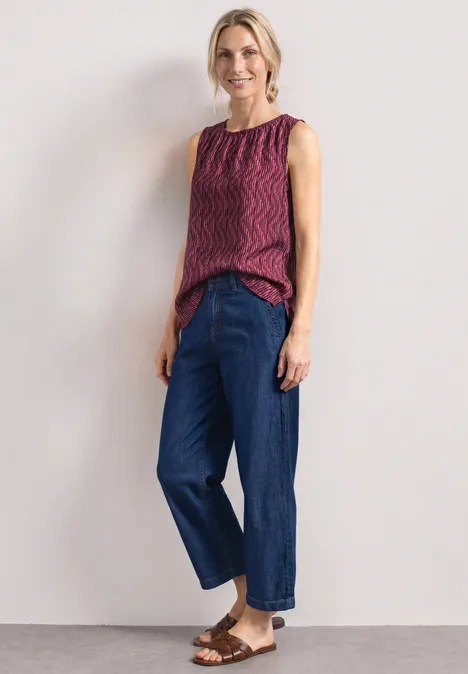 Femme en top rouge à motifs, jean bleu et sandales brunes devant un fond blanc.