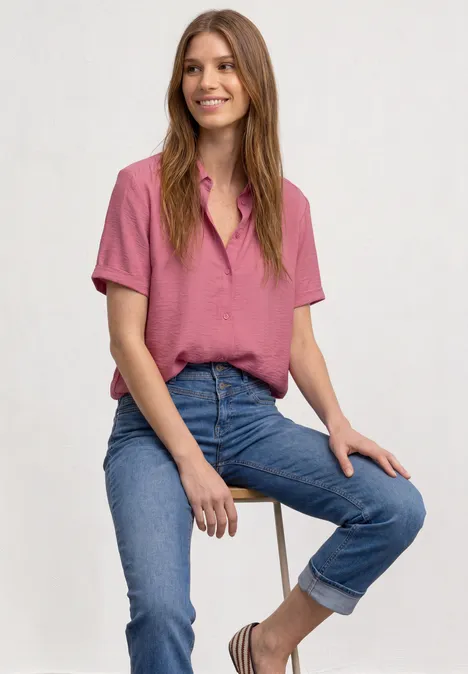 Een glimlachende vrouw zit op een kruk, draagt een roze blouse en opgerolde blauwe spijkerbroek.