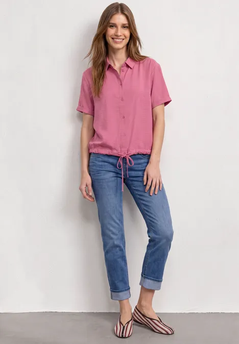 Femme souriante en chemise rose, jean retroussé et sandales, posant devant un mur blanc.