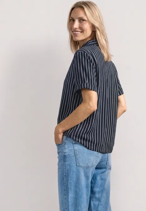 Person mit blau-weiß gestreiftem Hemd und Jeans, Hände in den Taschen, halb seitlich von hinten.