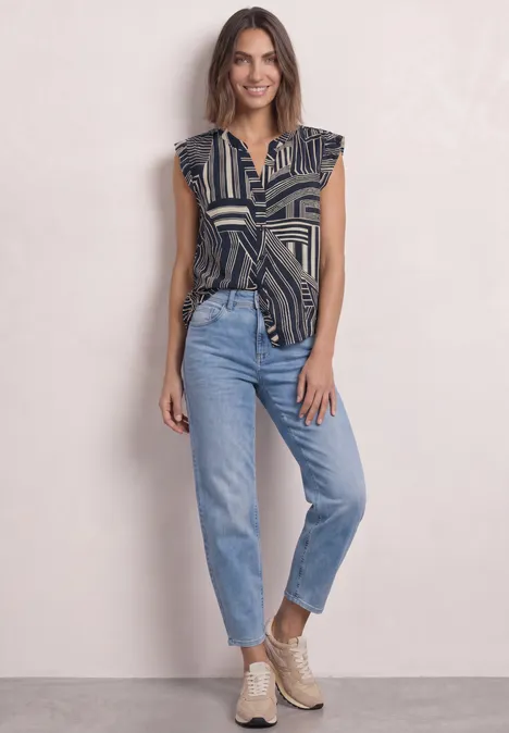 Eine Frau in gemusterter Bluse, hochgekrempelten Jeans und beigen Stiefeletten lächelt vor hellem Hintergrund.