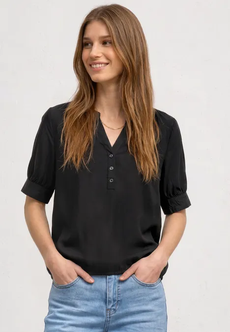 Kurzarm Bluse mit Split Neck und Elastiksaum Black Person mit schwarzem Oberteil und Jeans steht locker mit Händen in den Taschen vor neutralem Hintergrund.