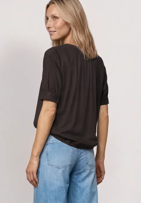 Blouse met korte mouwen, split in de hals en elastische zoom black coffee Frau steht mit Blick nach hinten, trägt schwarzes T-Shirt mit hochgekrempelten Ärmeln und helle Jeans.