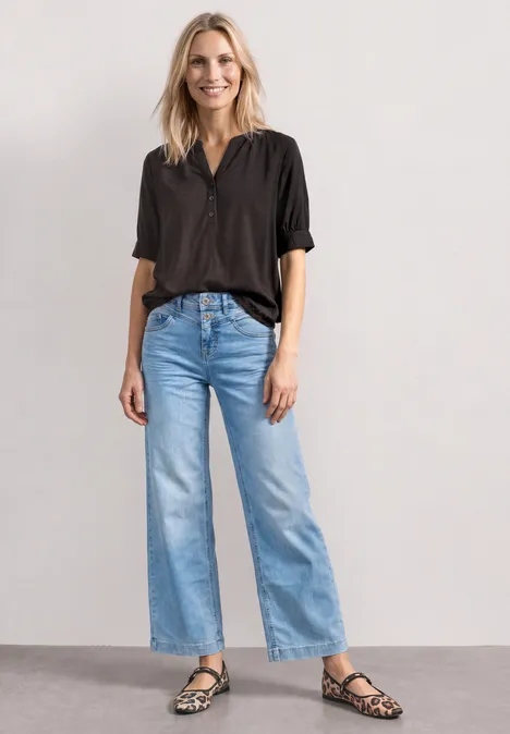 Blouse met korte mouwen, split in de hals en elastische zoom black coffee Eine blonde Frau in schwarzem Hemd, hellblauer Jeans mit weitem Bein und gemusterten Sandalen steht vor heller Wand.