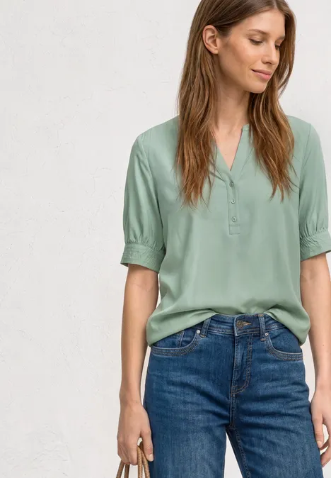Kurzarm Bluse mit Split Neck und Elastiksaum eucalyptus green Frau in grüner Bluse und blauen Jeans, steht und hält etwas in der rechten Hand.