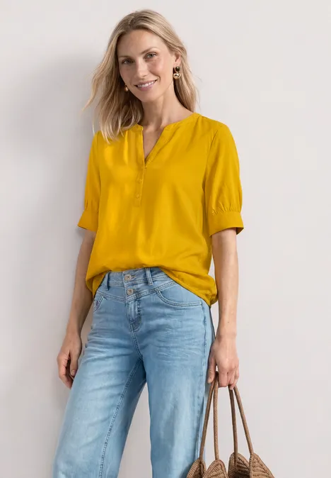 Blouse met korte mouwen, split in de hals en elastische zoom saffron yellow Frau in gelber Bluse und blauen Jeans trägt braune Handtasche vor weißem Hintergrund.