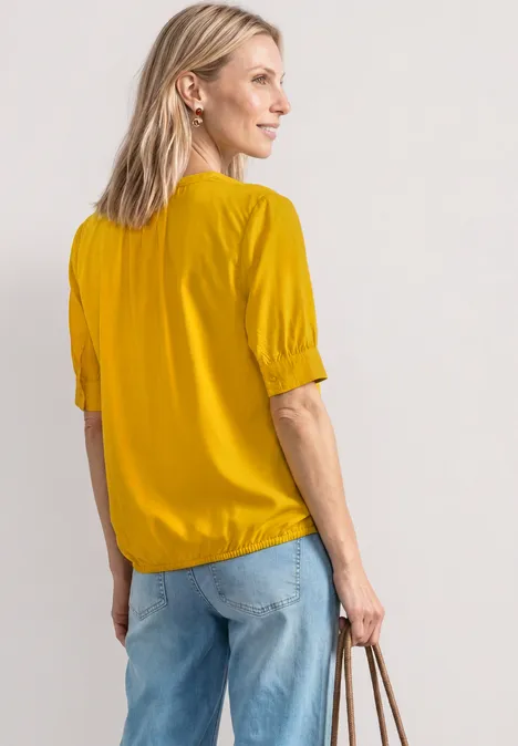 Blouse met korte mouwen, split in de hals en elastische zoom saffron yellow Eine Frau in gelber Bluse und Jeans von der Seite, sie hält eine Handtasche.