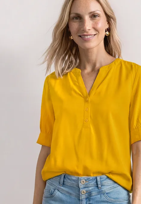 Blouse met korte mouwen, split in de hals en elastische zoom saffron yellow Eine Frau trägt eine senfgelbe Bluse mit V-Ausschnitt und halblangen Ärmeln, lässig in helle Jeans gesteckt.