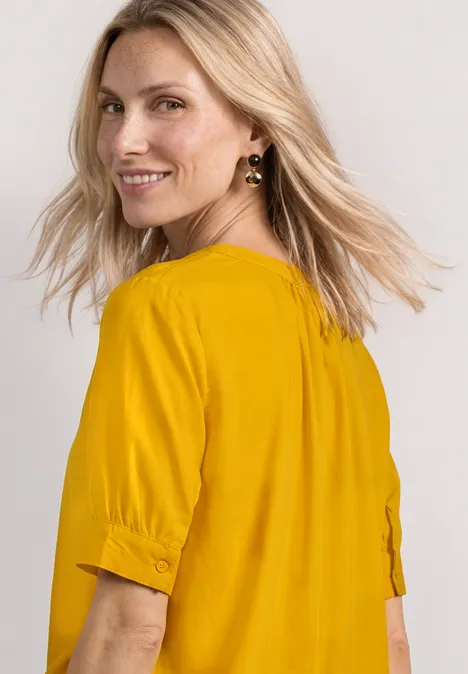 Blouse met korte mouwen, split in de hals en elastische zoom saffron yellow Frau mit blonden Haaren trägt gelbes Oberteil mit halblangen Ärmeln, seitlich fotografiert.