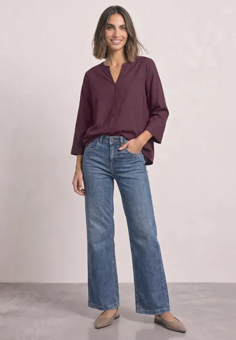 Femme portant un chemisier violet et un jean bleu évasé sur fond neutre.