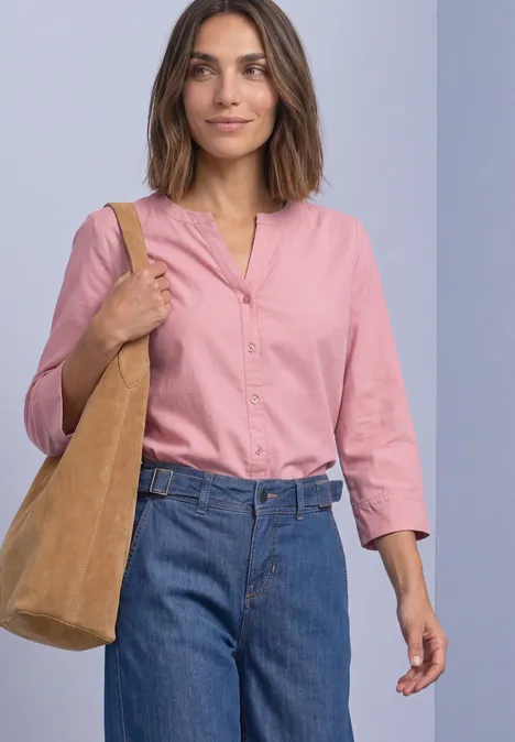 Chemisier à manches 3/4 et col fendu, en velours côtelé vintage blush Frau in rosa Bluse, blauen Jeans und beiger Schultertasche vor neutralem Hintergrund.