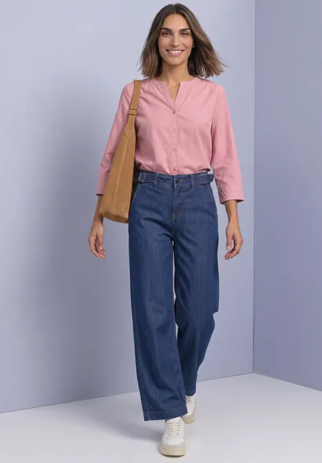 Chemisier à manches 3/4 et col fendu, en velours côtelé vintage blush Frau in blauer Jeans, rosa Bluse und weißen Sneakern mit Schultertasche geht vor hellviolettem Hintergrund.