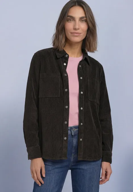 Frau vor neutralem Hintergrund trägt braune Cord-Überhemdjacke, rosa Top und Jeans.