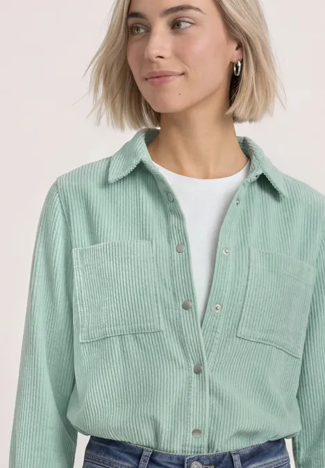 Frau mit kurzem blonden Haar trägt hellgrünes Kordhemd über weißem T-Shirt.