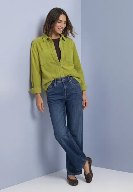 Langarm Hemdblsue aus Cord mit Knöpfen apple lime Frau lehnt an Wand, trägt grüne Bluse, dunkles Top, blaue Jeans und braune Schuhe.