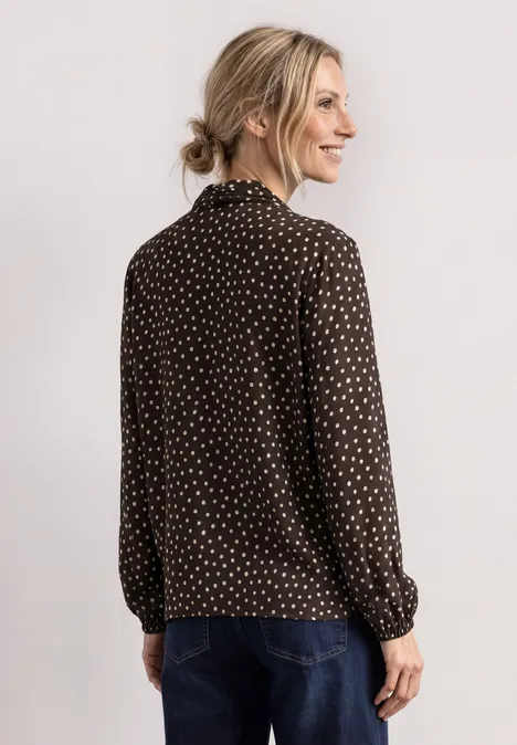 Frau im Profil trägt dunkle, gepunktete Bluse und Jeans, blondes Haar locker gebunden, vor hellem Hintergrund.
