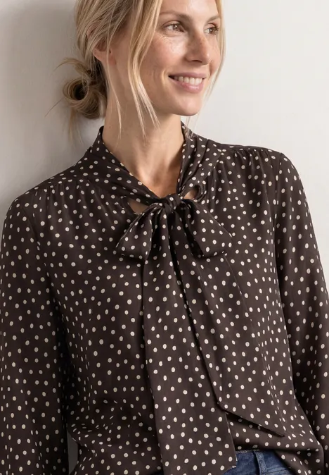 Blonde Frau in brauner Bluse mit weißem Herzmuster und Schleife am Kragen, lächelnd zur Seite blickend.