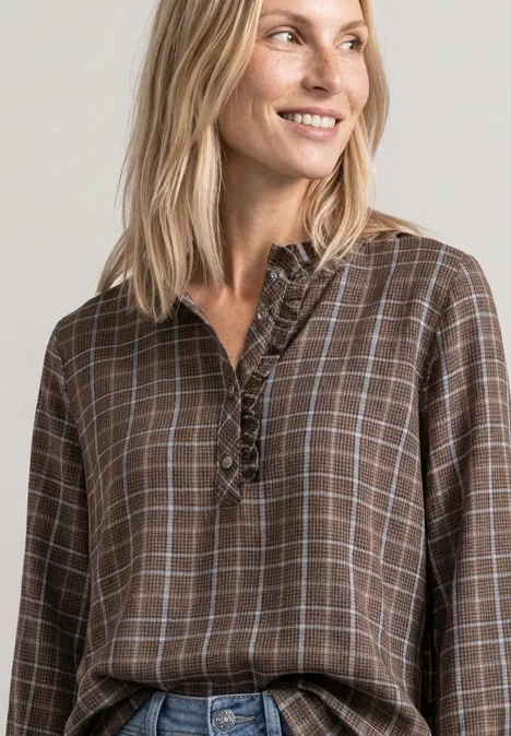 Blonde Frau lächelt seitlich, trägt braun-kariertes Blusenshirt mit Rüschen vor neutralem Hintergrund.