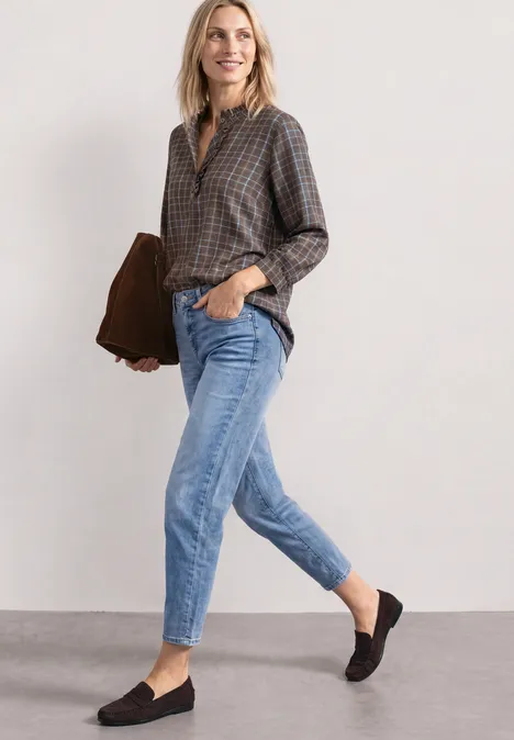 Eine lächelnde Frau in kariertem Hemd, Jeans und Mokassins hält eine braune Tasche.