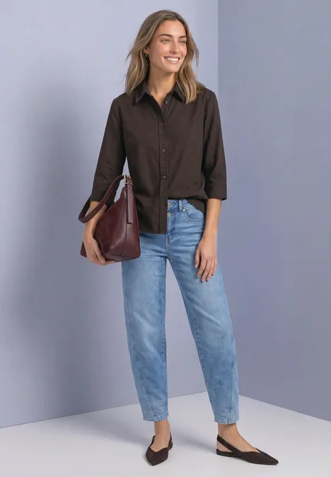 Eine Frau in braunem Hemd, hellblauen Jeans, schwarzen Slingback-Schuhen und großer bordeauxfarbener Tasche vor grauer Wand.