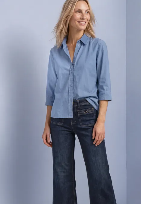 Dame trägt hellblaue Bluse und dunkle Jeans, lächelt vor hellem Hintergrund.