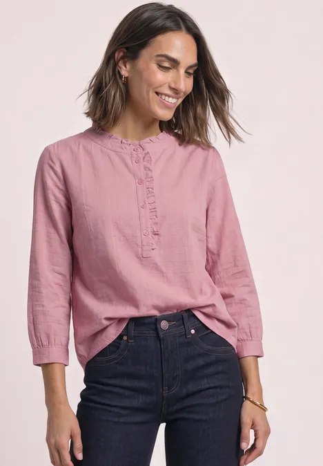 Frau in rosa Bluse und dunklen Jeans, lächelt vor hellem Hintergrund.