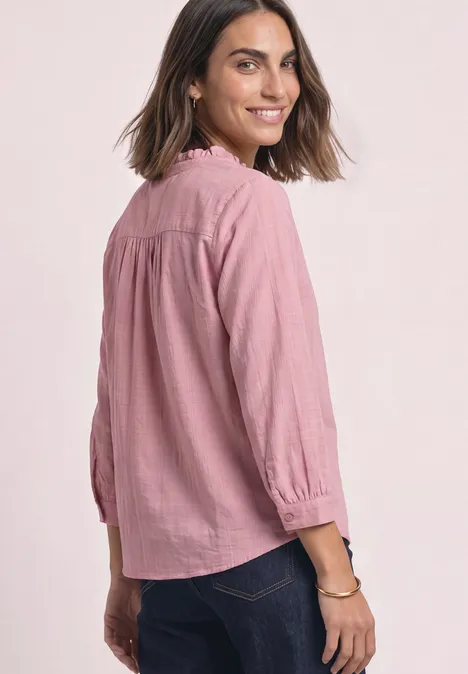 Frau in rosa Bluse mit 3/4-Ärmeln, Rückenansicht, lächelt, trägt dunkle Jeans.