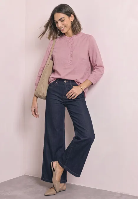 Frau in rosa Bluse, weiten dunklen Jeans und beigen Pumps, vor hellem Hintergrund lächelnd.