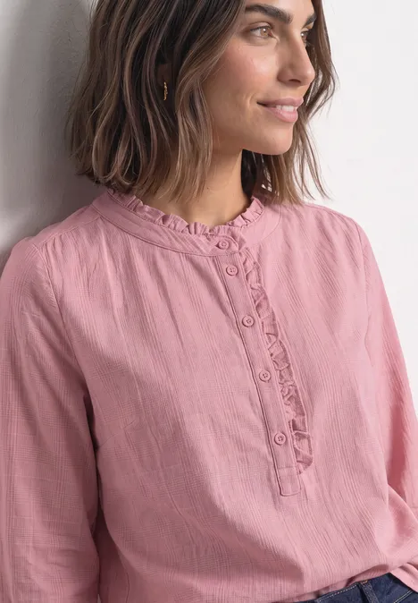 Eine Frau in rosa Bluse lehnt an einer Wand und blickt seitlich mit sanftem Lächeln.