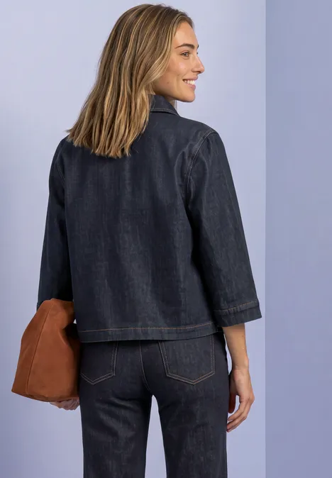 Frau von hinten in dunkelblauem Jeans-Outfit mit brauner Tasche vor hellem Hintergrund.