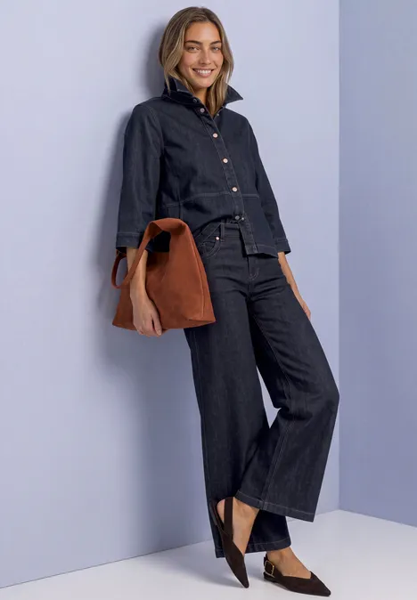 Frau in dunklem Denim-Outfit mit weiter Hose, Jeansjacke, schwarzen Schuhen und brauner Tasche vor hellem Hintergrund.