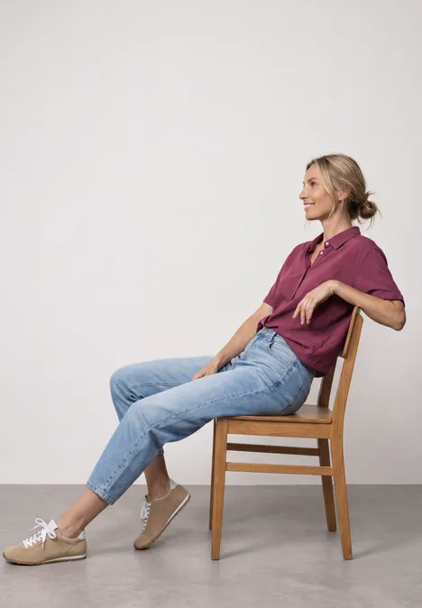 Femme assise de profil sur chaise en bois, détendue, vêtue d’un t-shirt violet, jean et baskets beiges.