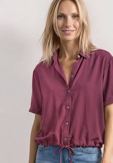 Femme portant une chemise manches courtes fuchsia à taille coulissante, souriante devant un fond clair.