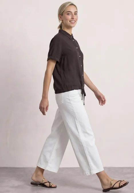 Overhemd met korte mouwen in effen kleur black coffee Een lachende vrouw in zwart shirt, witte broek en sandalen loopt.