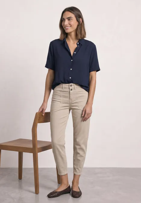Femme en chemise bleu marine à manches courtes, pantalon beige et mocassins, debout près d'une chaise sur fond clair.