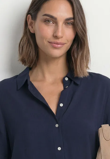 Portrait d'une femme aux cheveux mi-longs bruns portant un chemisier bleu marine devant un mur clair.