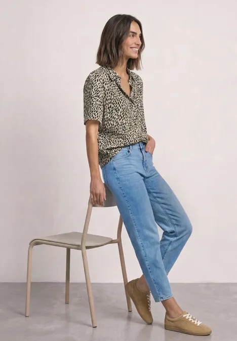 Overhemd met korte mouwen en print cotton beige Frau in Leopardenmuster-Bluse, blauen Jeans und beigen Schuhen lehnt lässig an einem Stuhl.