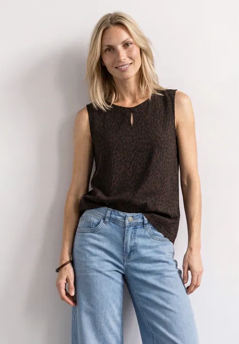 Lächelnde Frau im schwarzen Top und blauen Jeans lehnt entspannt an weißer Wand.