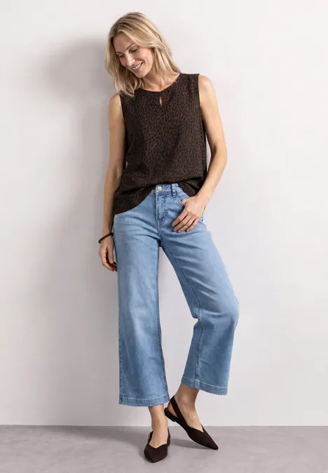 Vrouw in donkerbruin mouwloos topje, losse lichtblauwe wijde jeans en platte donkere schoenen.