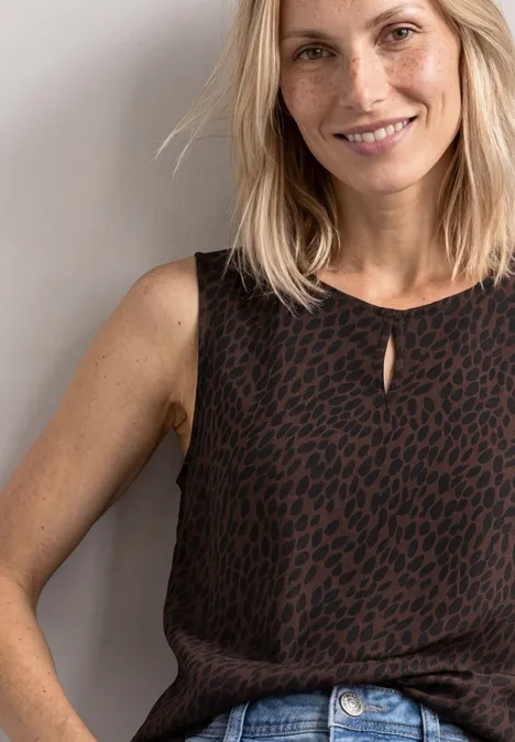 Vrouw met blond haar, gekleed in een bruin, motiefmouwloos top, glimlachend voor een lichte achtergrond.