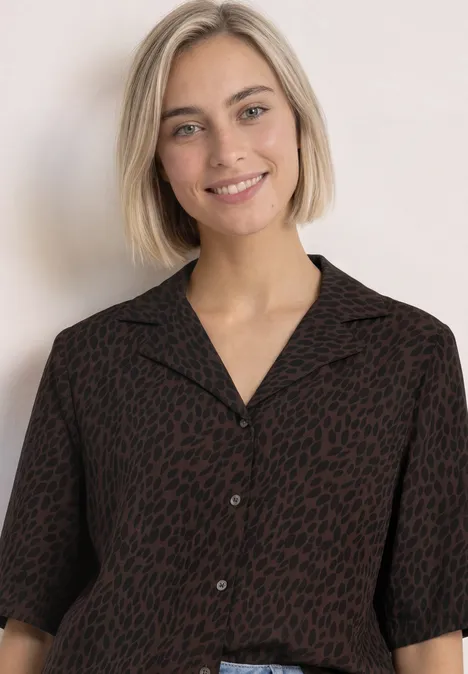 Junge Frau mit kurzem blonden Haar lächelt in einer dunkel gemusterten Bluse vor hellem Hintergrund.