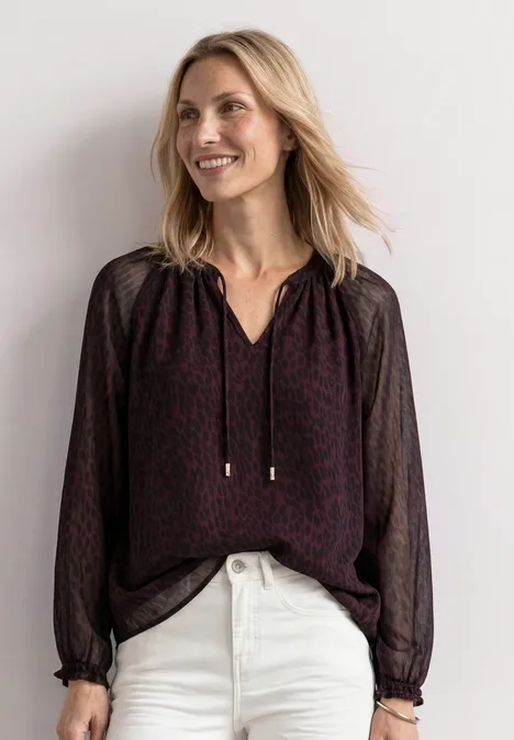 Eine lächelnde Frau trägt eine dunkle, gemusterte Bluse mit V-Ausschnitt und weiße Jeans.