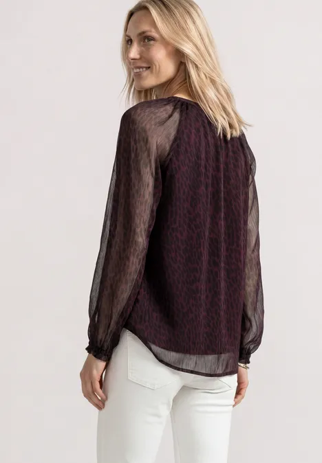 Raglanblouse met split in de hals en print rustic rouge Een vrouw draagt een bordeauxblouse met zwarte, doorschijnende mouwen en witte broek.
