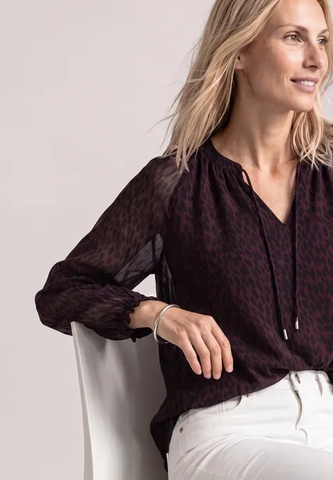 Raglanblouse met split in de hals en print rustic rouge Een blonde vrouw in een donker bloesje en lichte broek zit op een stoel en glimlacht.