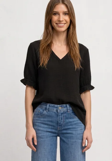 Kurzarm Bluse mit V-Neck und Rüschen Black Lächelnde Frau in schwarzem V-Ausschnitt-Shirt und blauen Jeans, steht mit Händen an den Hüften.