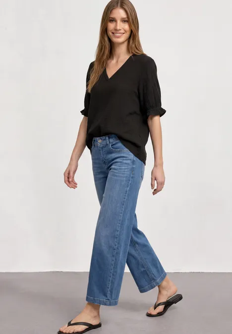 Kurzarm Bluse mit V-Neck und Rüschen Black Frau in schwarzem V-Neck-Oberteil, weiten Jeans und schwarzen Flip-Flops vor schlichtem Hintergrund.